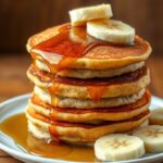 Delicious stack of banana pancakes, dripping with syrup and topped with banana slices.